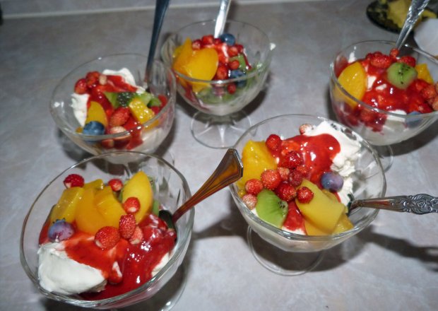 Fotografia przedstawiająca tęczowe lody z owocami i bitą śmietaną z polewą truskawkową